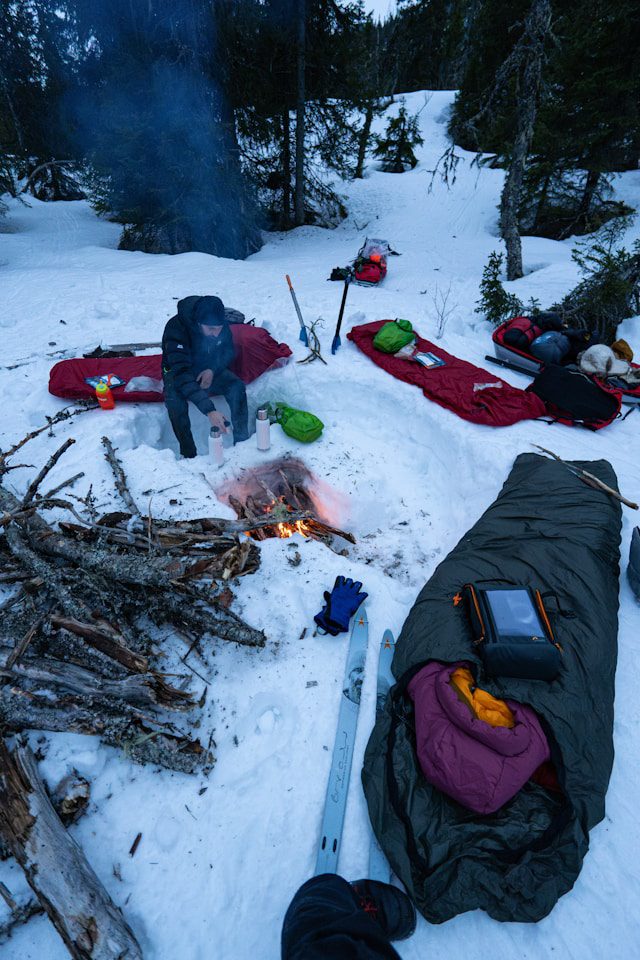 A sleeping bag, emergency kit, and skis near a fire in the snow.